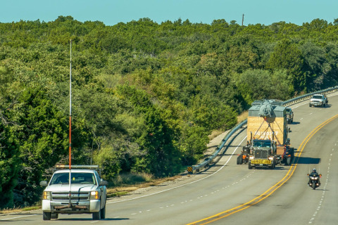 A high pole pilot car drives in front of a hydraulic platform trailer hauling an oversize load on the highway